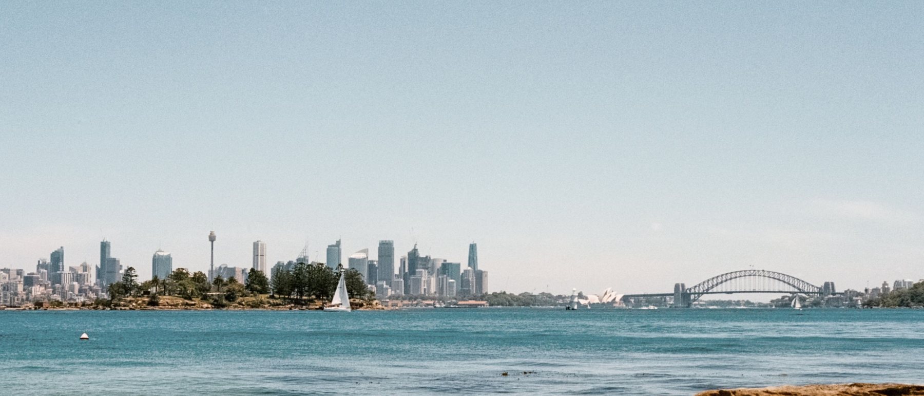 Sydney Harbour seen from a sandstone headland, the city skyline and the Harbour Bridge small on a distant shore under an open sky